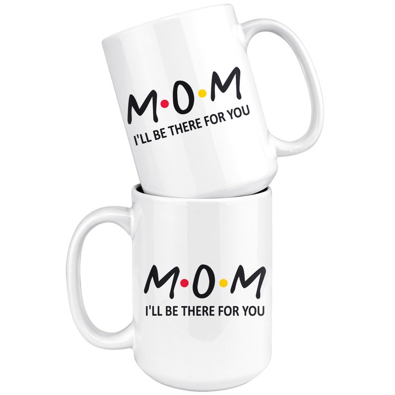 May include: Two white ceramic mugs with black text that reads "M.O.M. I'll be there for you". The "O" in "MOM" is a red dot and the "M" is a yellow dot.