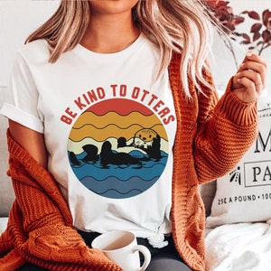 Pu&ograve; includere: Maglietta bianca con una grafica in stile retr&ograve; di lontre che nuotano nell'oceano. La grafica ha uno sfondo di tramonto rosso, arancione e giallo con il testo "Be Kind To Otters".