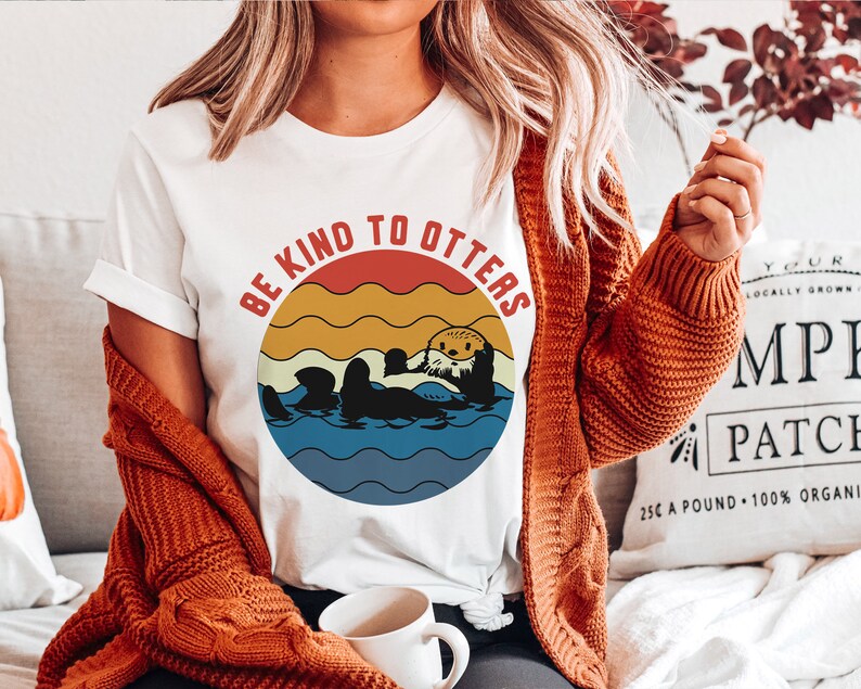 Pu&ograve; includere: Maglietta bianca con una grafica in stile retr&ograve; di lontre che nuotano nell'oceano. La grafica ha uno sfondo di tramonto rosso, arancione e giallo con il testo "Be Kind To Otters".