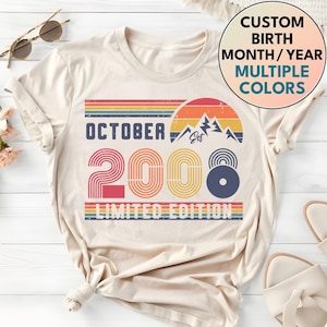 May include: A cream-colored t-shirt featuring a graphic with the text OCTOBER 2008 LIMITED EDITION. The design includes a retro sunset, mountain silhouettes, and striped lines in orange, yellow, and blue. A circular badge reads CUSTOM BIRTH MONTH/YEAR MULTIPLE COLORS.