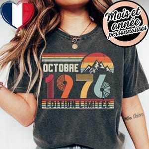 May include: A charcoal gray t-shirt featuring a graphic print with the text Octobre 1976 Edition Limitee. The design includes a sunset motif with horizontal stripes in orange, yellow, and blue, along with a mountain silhouette and the word Est.