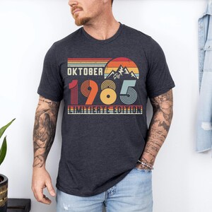 May include: A charcoal gray short-sleeve t-shirt featuring a retro-style graphic with a sunset, mountains, and the text "OKTOBER 1985 LIMITIERTE EDITION". The design uses horizontal stripes and circular patterns in shades of red, orange, and blue.