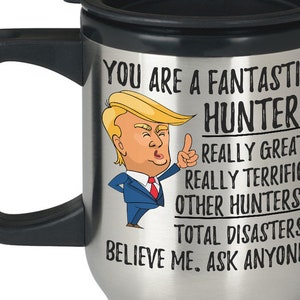 May include: A silver travel mug with a black handle and lid. The mug features a cartoon image of a person with blonde hair and a black suit. The text on the mug reads "You are a fantastic hunter. Really great. Really terrific. Other hunters? Total disasters! Believe me. Ask anyone."