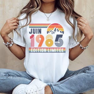 May include: A white t-shirt featuring a retro-style graphic with the text JUIN 1985 &Eacute;DITION LIMIT&Eacute;E. The design includes a sunset, mountain peaks, and horizontal stripes in shades of red, orange, yellow, blue, and green, with a distressed texture.