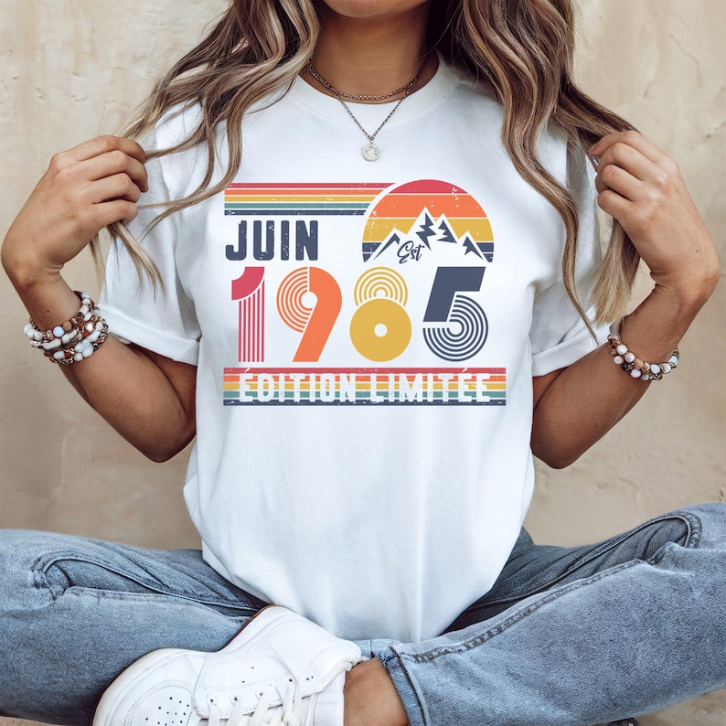 May include: A white t-shirt featuring a retro-style graphic with the text JUIN 1985 &Eacute;DITION LIMIT&Eacute;E. The design includes a sunset, mountain peaks, and horizontal stripes in shades of red, orange, yellow, blue, and green, with a distressed texture.