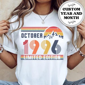May include: A white t-shirt featuring a retro-style graphic with the text OCTOBER 1996 LIMITED EDITION. The design includes a mountain silhouette, a sunset motif with horizontal stripes in muted colors, and the word Est in script lettering.