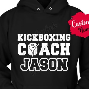 May include: Black hooded sweatshirt with white text that reads "Kickboxing Coach" and a silhouette of a person kickboxing. The text "Jason" is also in white below the main text. There is a red circle with white text that reads "Custom Name" in the upper right corner of the image.
