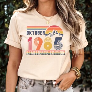 May include: A cream-colored short-sleeve t-shirt featuring a graphic print with the text "OKTOBER 1985 LIMITIERTE EDITION". The design includes a retro-style sunset, mountain silhouettes, and striped patterns in shades of red, orange, yellow, and blue.