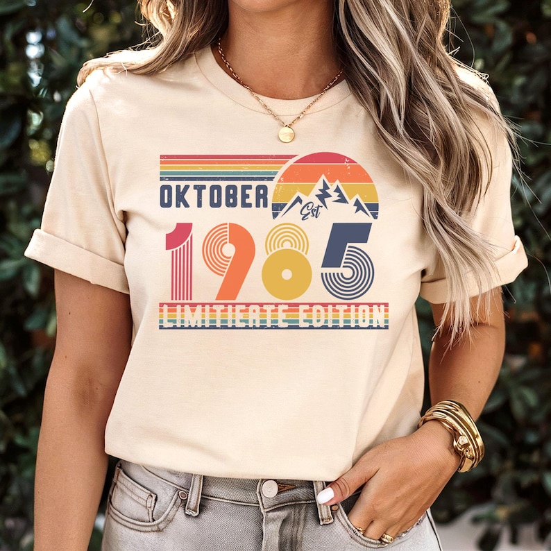 May include: A cream-colored short-sleeve t-shirt featuring a graphic print with the text "OKTOBER 1985 LIMITIERTE EDITION". The design includes a retro-style sunset, mountain silhouettes, and striped patterns in shades of red, orange, yellow, and blue.