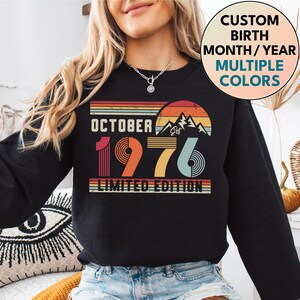 May include: A black long-sleeve sweatshirt featuring a graphic print with the text OCTOBER 1976 LIMITED EDITION. The design includes a retro-style sunset, mountain silhouette, and horizontal stripes in shades of red, orange, yellow, and teal blue.