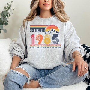 May include: A light gray crewneck sweatshirt featuring a retro-style graphic with the text SEPTEMBER 1985 LIMITIERTE EDITION. The design includes a sunset, mountain range, and horizontal stripes in shades of red, orange, yellow, and blue.