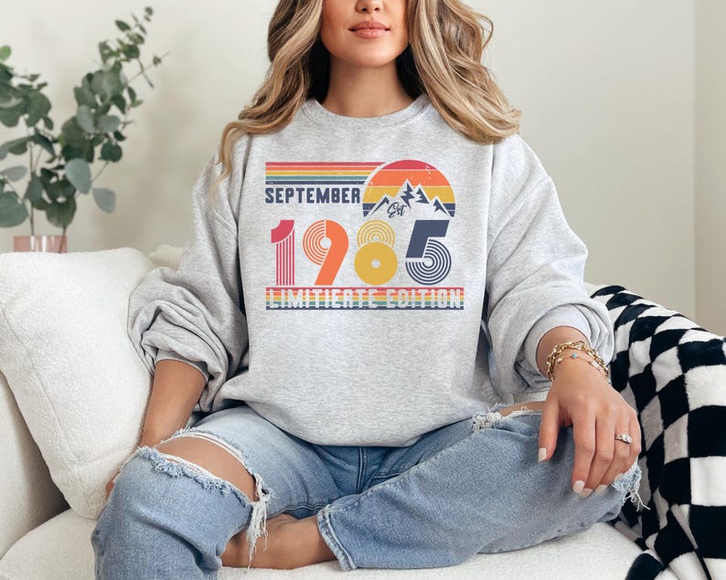 May include: A light gray crewneck sweatshirt featuring a retro-style graphic with the text SEPTEMBER 1985 LIMITIERTE EDITION. The design includes a sunset, mountain range, and horizontal stripes in shades of red, orange, yellow, and blue.