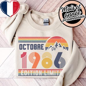 May include: A cream-colored crewneck sweatshirt featuring a graphic print with the text OCTOBRE 1986 EDITION LIMITEE. The design includes a retro-style sunset, mountain peaks, and horizontal stripes in shades of red, orange, yellow, and blue.