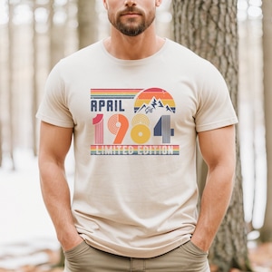 May include: A beige short-sleeve t-shirt featuring a graphic print with the text "APRIL 1984 LIMITED EDITION". The design includes a retro-style sunset, mountain silhouettes, and horizontal stripes in shades of red, orange, yellow, and blue.