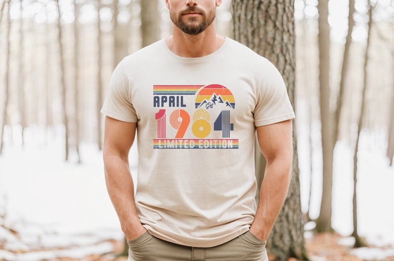 May include: A beige short-sleeve t-shirt featuring a graphic print with the text "APRIL 1984 LIMITED EDITION". The design includes a retro-style sunset, mountain silhouettes, and horizontal stripes in shades of red, orange, yellow, and blue.