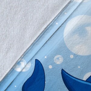 May include: A blue and white fleece blanket with a pattern of blue whales and white bubbles. The blanket is folded in half, showing the white fleece on one side and the blue and white patterned fleece on the other side.