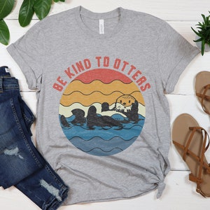 Pu&ograve; includere: Una maglietta grigia con una grafica in stile vintage di una lontra in acqua. La grafica ha uno sfondo di tramonto con il testo "Be Kind to Otters".