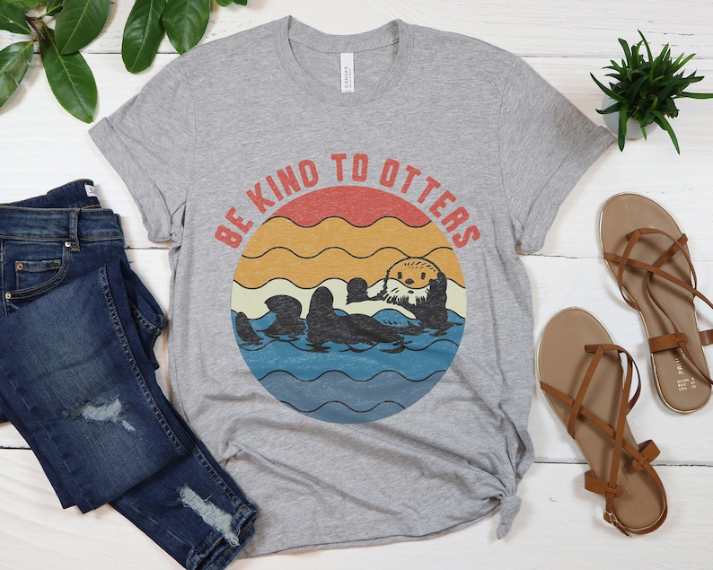 Pu&ograve; includere: Una maglietta grigia con una grafica in stile vintage di una lontra in acqua. La grafica ha uno sfondo di tramonto con il testo "Be Kind to Otters".
