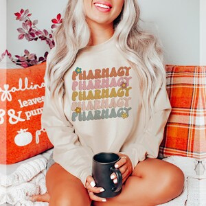 May include: A beige sweatshirt with the word "Pharmacy" repeated five times in a retro font with flowers. The sweatshirt is worn by a person sitting on a couch with a mug in their hand.