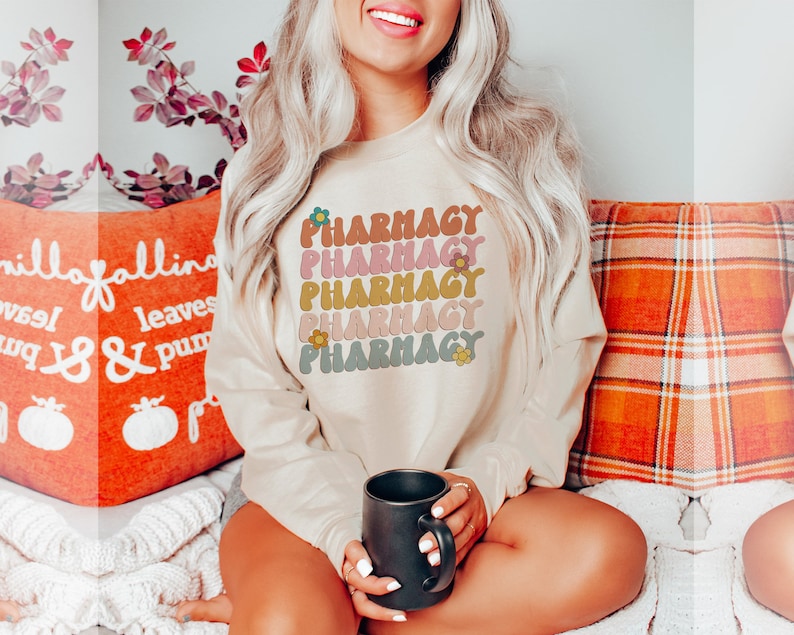 May include: A beige sweatshirt with the word "Pharmacy" repeated five times in a retro font with flowers. The sweatshirt is worn by a person sitting on a couch with a mug in their hand.