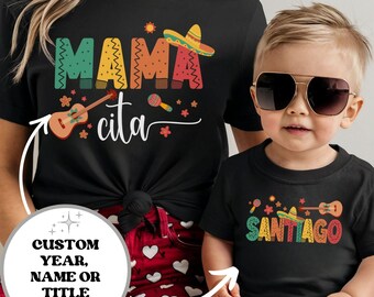 May include: Matching black t-shirts for a parent and child. The adult shirt says "Mamacita" in colorful letters with a guitar, maracas, and a sombrero. The child's shirt says "Santiago" in colorful letters with a guitar, maracas, and a sombrero.