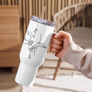 May include: White insulated tumbler with a handle, a clear lid, and a straw. The tumbler features a black line drawing of hands holding each other, with names like "Papi" and "Lucie" written next to them. The tumbler is held by a person.