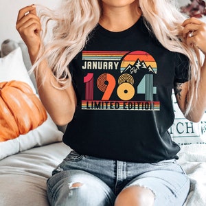 May include: A black t-shirt featuring a retro-style graphic with a sunset, mountains, and the text JANUARY 1984 LIMITED EDITION. The design uses horizontal stripes in orange, yellow, and teal, with the year 1984 displayed in large, colorful block lettering.