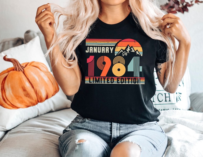 May include: A black t-shirt featuring a retro-style graphic with a sunset, mountains, and the text JANUARY 1984 LIMITED EDITION. The design uses horizontal stripes in orange, yellow, and teal, with the year 1984 displayed in large, colorful block lettering.