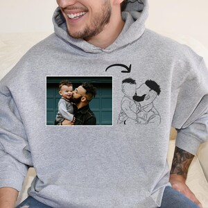 Könnte beinhalten: Ein grauer Hoodie mit einer Schwarz-Weiß-Zeichnung eines Vaters und Sohnes auf der Vorderseite. Die Zeichnung ist eine stilisierte Version eines Fotos eines Vaters und Sohnes, die sich auf der Vorderseite des Hoodies küssen.