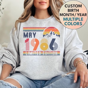 Op de afbeelding: Een lichtgrijs sweatshirt met een retro design. Het ontwerp bevat de tekst "MAY 1986 LIMITED EDITION", een regenboog en een berg-afbeelding. Een cirkel toont "CUSTOM BIRTH MONTH/YEAR MULTIPLE COLORS".