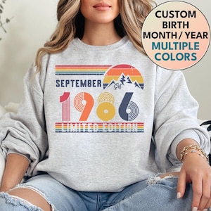 Op de afbeelding: Een lichtgrijs sweatshirt met ronde hals en een grafische print met de tekst SEPTEMBER 1986 LIMITED EDITION. Het ontwerp bevat een retro zonsondergang, bergtoppen en horizontale strepen in de kleuren rood, oranje, geel en blauw.