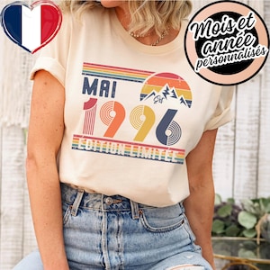 May include: Cream-coloured t-shirt featuring the text "MAI 1996 EDITION LIMITEE" in a retro style. The design incorporates a rainbow, mountains, and the year 1996 in red, orange, and blue. The shirt is worn with blue denim shorts.