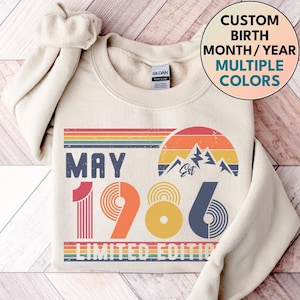 Puede incluir: Sudadera crema con un diseño retro que presenta el texto "MAY 1986" en azul, naranja y marino. Un arcoíris y un gráfico de montaña están encima del año. El texto "LIMITED EDITION" está en la parte inferior. La imagen también incluye el texto "CUSTOM BIRTH MONTH/YEAR MULTIPLE COLORS".