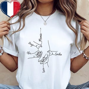 Puede incluir: Camiseta blanca con un dibujo lineal negro de manos entrelazadas. Los nombres "Hugo", "Alex", "Lucie" y "Sacha" están escritos cerca de las manos. La palabra "Mamie" está escrita encima de las manos. Un corazón con la bandera francesa está en la esquina superior izquierda.