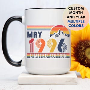 May include: White ceramic mug with a black handle and rim. The mug features a retro design with the text "MAY 1996 LIMITED EDITION" in colorful stripes. A circular graphic with mountains and the text "CUSTOM MONTH AND YEAR MULTIPLE COLORS" is also visible.