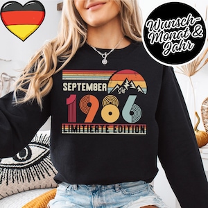 May include: A black long-sleeve sweatshirt featuring a retro-style graphic with a sunset, mountains, and the text "SEPTEMBER 1986 LIMITIERTE EDITION". The design uses horizontal stripes in orange, yellow, and red tones above and below the text.