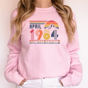 May include: A light pink sweatshirt with the text "APRIL 1984 LIMITED EDITION" in a retro design. The graphic includes a mountain scene and rainbow stripes, perfect for a birthday or anniversary gift.
