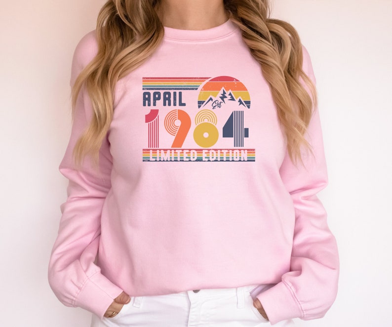 May include: A light pink sweatshirt with the text "APRIL 1984 LIMITED EDITION" in a retro design. The graphic includes a mountain scene and rainbow stripes, perfect for a birthday or anniversary gift.