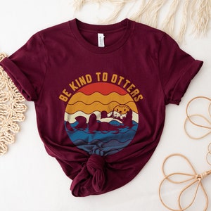Pu&ograve; includere: Una maglietta color marsala con una grafica vintage di una lontra in acqua. La grafica dice "Be Kind to Otters".