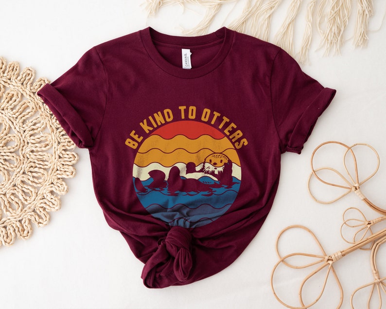 Pu&ograve; includere: Una maglietta color marsala con una grafica vintage di una lontra in acqua. La grafica dice "Be Kind to Otters".