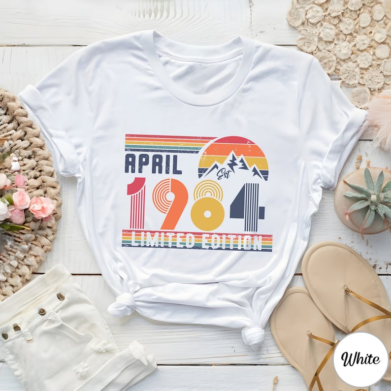 May include: A white short-sleeve t-shirt featuring a retro-style graphic with the text "APRIL 1984 LIMITED EDITION". The design includes a sunset, mountain peaks, and horizontal stripes in red, orange, yellow, and blue, with a distressed texture.