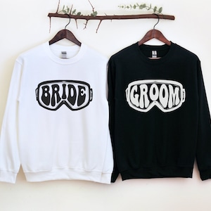 May include: Two white and black sweatshirts with the words "Bride" and "Groom" printed on them in a retro ski goggle design.