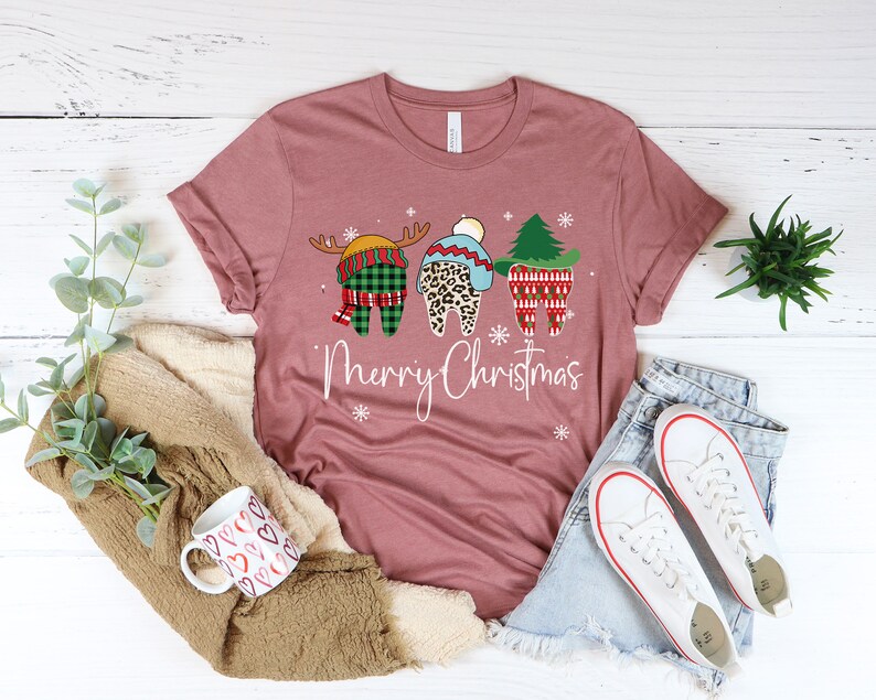Op de afbeelding: Mauvekleurig T-shirt met een "Merry Christmas"-afbeelding met drie tanden gekleed in feestelijke kleding. E&eacute;n tand is een rendier, een andere heeft een luipaardprint en de derde is een kerstboom. Witte sneeuwvlokken zijn verspreid rond de tekst.