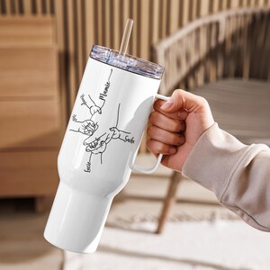 May include: White insulated tumbler with a handle and a clear lid, held by a person. The tumbler features a black line drawing of hands and the names "Hugo", "Alex", "Lucie", "Sacha", and "Mamie". A clear straw is inserted in the lid.