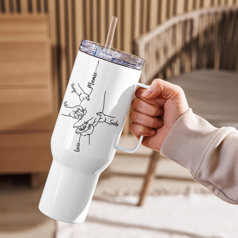 May include: White insulated tumbler with a handle and a clear lid, held by a person. The tumbler features a black line drawing of hands and the names "Hugo", "Alex", "Lucie", "Sacha", and "Mamie". A clear straw is inserted in the lid.