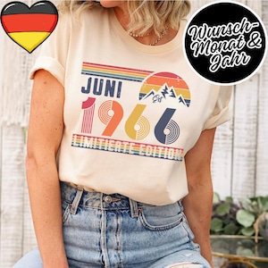 May include: Cream-colored t-shirt with the text "JUNI 1966 LIMITED EDITION" in a retro design. The design includes a rainbow, mountain graphic, and the word "EST". A German flag heart is in the upper left corner.