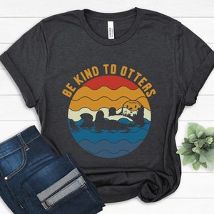 Pu&ograve; includere: Una maglietta grigio scuro con una grafica in stile vintage che dice "Be Kind to Otters". La grafica presenta una lontra dei cartoni animati che nuota in un oceano blu con un tramonto alle sue spalle.
