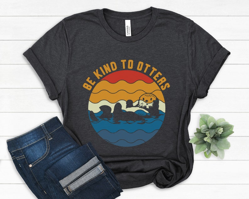 Pu&ograve; includere: Una maglietta grigio scuro con una grafica in stile vintage che dice "Be Kind to Otters". La grafica presenta una lontra dei cartoni animati che nuota in un oceano blu con un tramonto alle sue spalle.