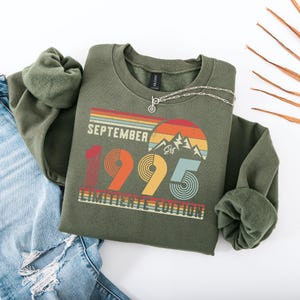 K&ouml;nnte beinhalten: Ein olivgr&uuml;nes Sweatshirt mit dem Text "September 1995 Limited Edition" in einem Retro-Design. Die Grafik enth&auml;lt eine Bergkette und ein Farbschema im Sonnenuntergangsstil. Eine silberne Halskette liegt auf dem Sweatshirt.