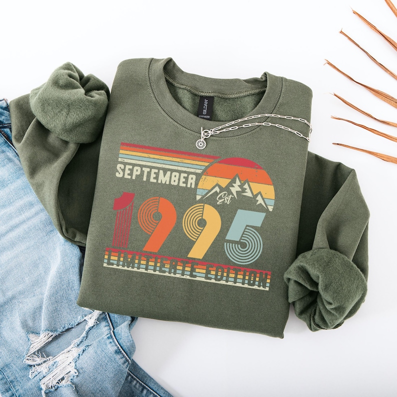 K&ouml;nnte beinhalten: Ein olivgr&uuml;nes Sweatshirt mit dem Text "September 1995 Limited Edition" in einem Retro-Design. Die Grafik enth&auml;lt eine Bergkette und ein Farbschema im Sonnenuntergangsstil. Eine silberne Halskette liegt auf dem Sweatshirt.